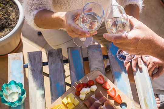 close up of two hands, a pair, from adult aged caucasian people do cheers with white wine and eat some fruits and snacks on a wood table outdoor under a sunny day nice weather. happiness 