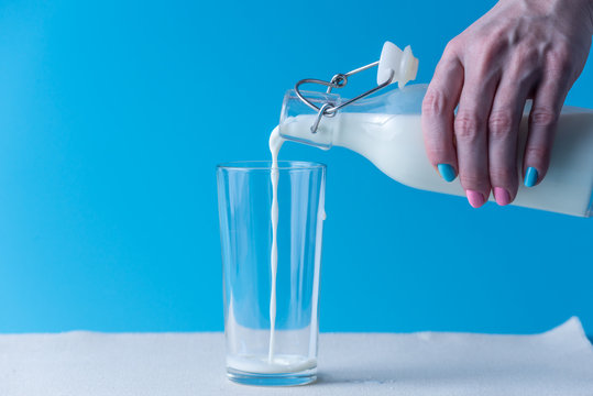 Woman's hand pours fresh milk from a glass bottle into a glass on a blue background. Healthy dairy products with calcium