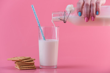 Woman's hand pours fresh milk from a glass bottle into a glass and cookies on a pink background. Healthy dairy products