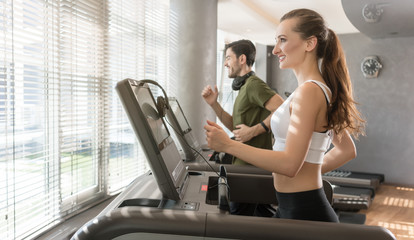 Fototapeta premium Happy young woman listening to music while running beside a handsome man on a modern treadmill with touch screen and headphones