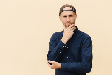 Confident European male holds chin, dressed in shirt and cap, has serious expression, poses against beige background. Handsome man thinks about something. People, lifestyle, masculinity concept