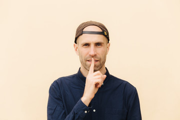 Horizontal shot of handsome young male model keeps fore finger on lips, asks to be quiet, wears cap...
