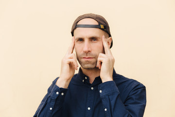 Indoor shot of attractive male keeps fingers on temples, tries to concentrate on some problem, recollects important information in mind, poses against blank studio wall. People and facial expressions