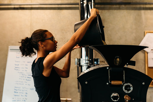 Young businesswoman pouring coffee beans into a roasting machine at her coffee shop. 