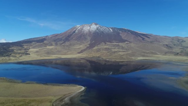 Tromen volcano in national park, Patagonia. Aerial scene moving backwards. Mountain with bed of lava and snow. Tromen lagoon with refletion of volcano. Wilde lonely landscape