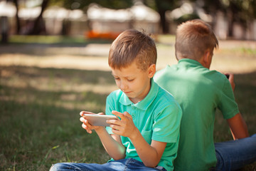 Adorable boy sitting on the grass in the park and playing with smartphone. Child learning how to use smartphone