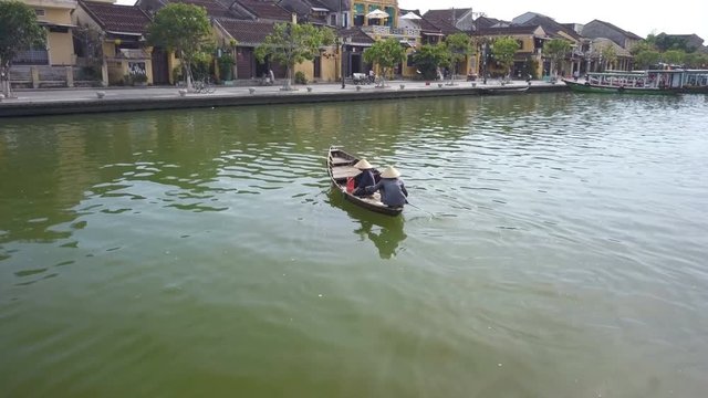 Drone follow shot of an elderly Vietnamese boat taxi woman paddling across a small river in Hoi An with a pedestrian, both wearing non la rice farmer hats. Stunning backdrop of ancient architecture
