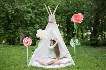 Little girl lying and playing in a tent, children's house wigwam in park. Happy mothers day. Child with big donuts and heart-shaped pillows. Summer, childhood concept. © MartaKlos