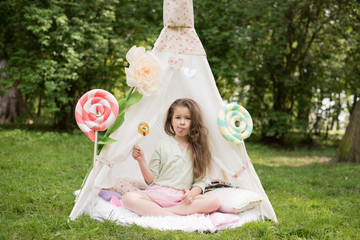 Little girl lying and playing in a tent, children's house wigwam in park. Happy mothers day. Child with big donuts and heart-shaped pillows. Summer, childhood concept. © MartaKlos