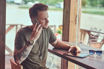 Handsome stylish hipster sits at a table in a roadside cafe, talking on a smartphone.