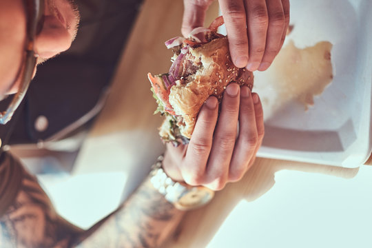Portrait of a hungry hipster guy with a stylish haircut and beard sits at a table, decided to dine at a roadside cafe, eating a hamburger. - Powered by Adobe