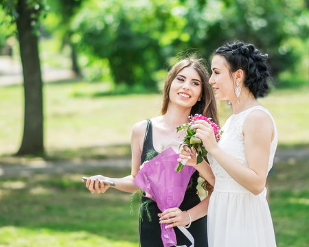 Portrait of two young pretty women friends talking in green summer park. Pretty females bride and bridesmaid smiling and chatting