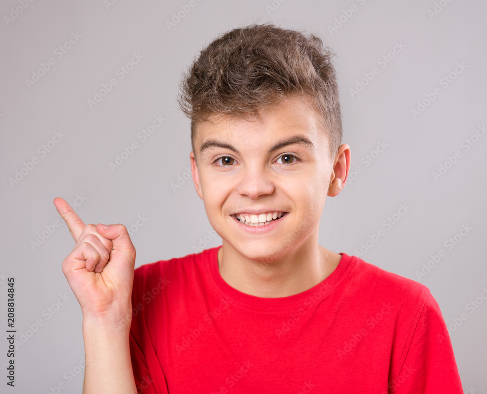 Wall mural Emotional portrait of excited teen boy. Funny cute surprised child 14 year old looking at camera. Happy teenager pointing fingers up, on gray background.