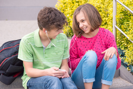 Happy teenage boy and girl with headphones are using gadget, talking and smiling while sitting on the stairs outdoors. Young sister and brother teens playing on mobile phone and listening to music.