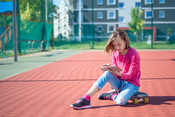 Happy teenage girl with headphones are using gadget, smiling while sitting on the playground outdoors. Young student teen with a skateboard playing on tablet pc, listening to music or watches video.
