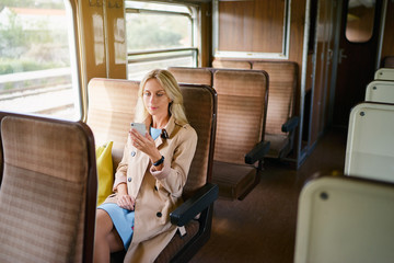 Enjoying trip. Young pretty woman traveling by the train sitting near the window using smartphone.