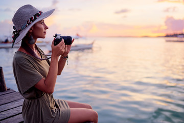 Photography and travel. Young woman in hat holding camera sitting on wooden fishing pier with...