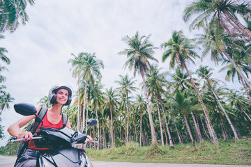 Tropical travel and transport. Young beautiful woman in helmet riding scooter on the road with palm trees.