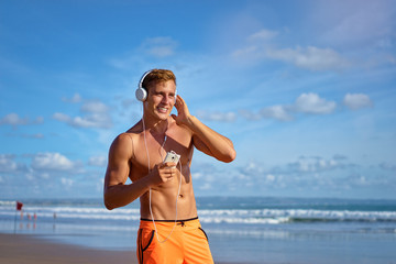 Enjoying music. Sports lifestyle. Happy young man in headphones walking on the sea shore.
