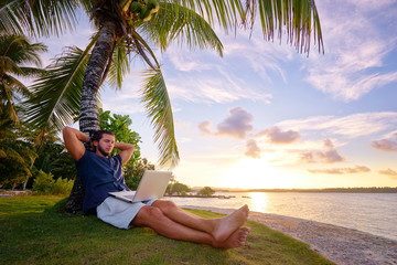 Work and vacation. Young man working on laptop computer on the tropical beach under the palm tree.