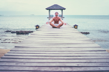 Yoga near the sea. Young man in lotus pose sitting on wooden pier. Concept of pray and meditation.
