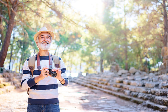 Enjoying travel. Senior smiling man with backpack holding camera on ancient sightseeing background.