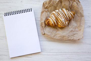 Freshly baked croissant with blank notebook on white wooden table, top view