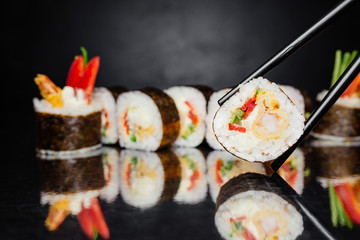 Chopsticks holding roll Philadelphia with tiger prawn on black background made of Seaweed, marinated rice, cheese Philadelphia, bell pepper, cucumber. Sushi menu. Japanese food. Free-space. Close-up.