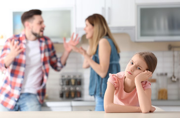 Little unhappy girl sitting at table while parents arguing on kitchen