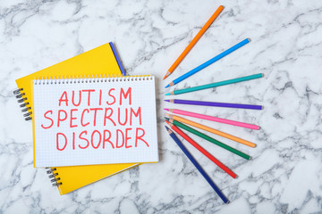 Notebook with words AUTISM SPECTRUM DISORDER and colorful pencils on marble background, top view