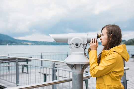 Girl in a yellow raincoat looking through coin-operated telescope at the opposite bank of the lake in Alps