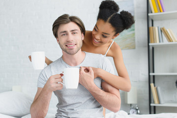 portrait of smiling multiracial couple with cups of coffee in bed in morning