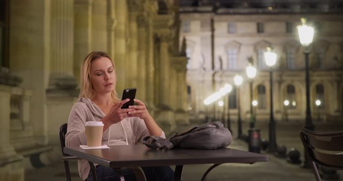 White lady sitting at table outside at night taking selfie by European building, Caucasian woman sitting alone at table using phone to take selfie photo outdoors at night, 4k
