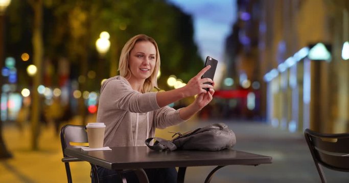 Peppy female taking selfie on phone while sitting at table on Champs-Elysees, Pretty young Caucasian lady taking selfie to send to friend in Paris, France in the evening, 4k