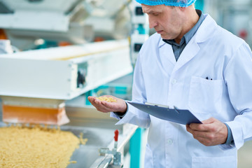 Mid section portrait of senior factory worker checking production process standing by conveyor belt holding handful of dry macaroni, copy space