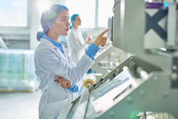 Fototapeta na wymiar Side view portrait of young female worker wearing lab coat and hair cover standing by power units pressing buttons on control panel in clean production workshop.
