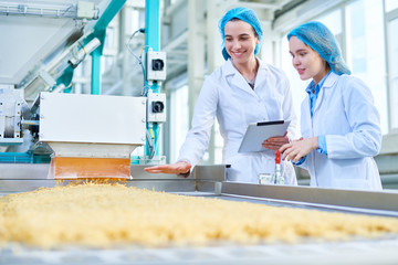 Waist up portrait of  two young female workers wearing lab coats standing by  conveyor line with macaroni  in clean production workshop, copy space