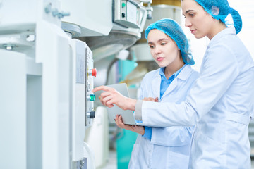 Side view portrait of  two young female workers wearing lab coats setting up power units and pushing buttons on control panel in clean production workshop, copy space