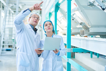 Portrait of senior factory worker wearing lab coat explaining rules to female trainee and pointing up while standing in clean production workshop of modern plant, copy space
