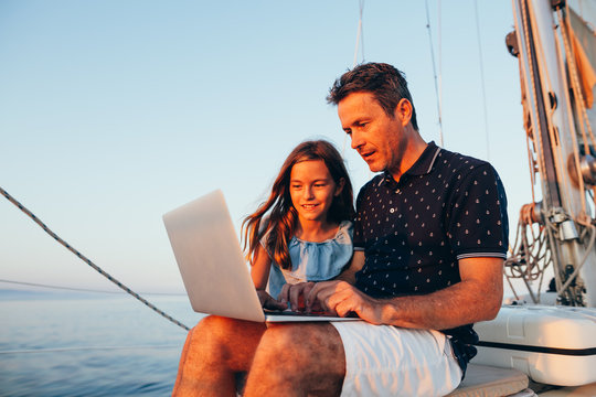 Father and daughter using laptop on a sailboat at sunset.