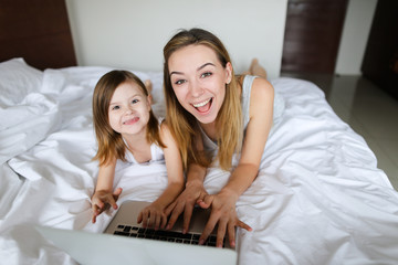 Happy blonde female person using laptop with little daughter and lying in bed. Concept of modern...