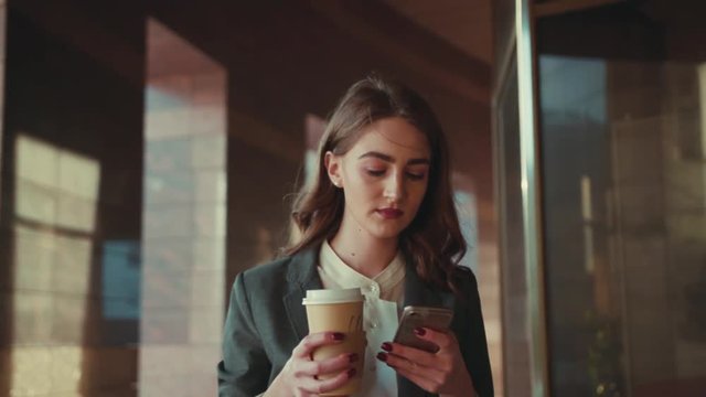 Attractive young woman in a trendy outfit walking along the modern shopping center, using her device and holding coffee. Modern active lifestyle, businesswoman, coffee break. Female portrait, sun lens