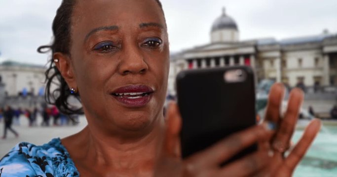 Happy black woman looks at pictures of her grandchildren on smartphone, Older African American woman in Trafalgar Square uses her mobile phone, 4k
