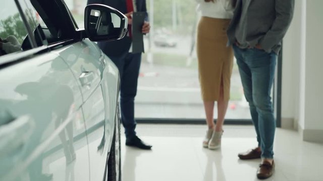 Low shot of prosperous people young couple buying new car in dealership and talking to helpful manager in suit. Shiny luxurious car is in foreground, large window in background.