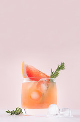 Fresh grapefruit juice with ice, piece and green rosemary closeup on soft white wood table and pink background, vertical.