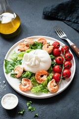 Salad with burrata cheese, shrimps, lettuce and tomatoes on a white plate on dark stone table. Healthy italian salad