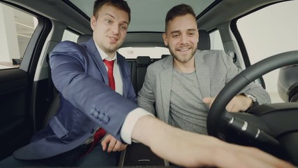 Car dealer is giving key fob to cheerful male buyer, men are shaking hands, smiling and talking sitting inside automobile after test drive. Buying transport and people concept.
