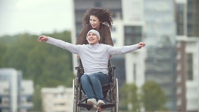 Happy disabled man in a wheelchair with happy young woman running at the city street