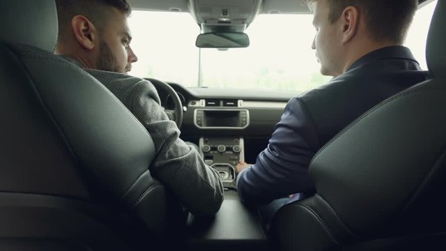 Back view of two men car dealer and customer talking discussing automobile model while sitting on front seats in beautiful auto. Leather seats and electronic board are visible.