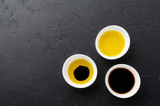 Soy sauce, olive oil and balsamic sauce in white ceramic bowls on black stone or concrete background.